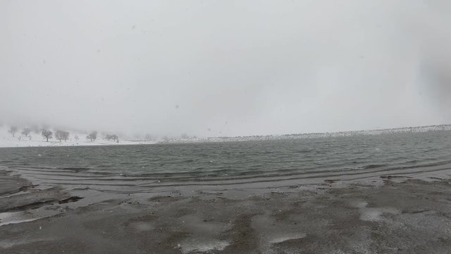 Gentle ripples reaching frozen banks of lake with fresh snow falling, Low Angle