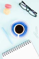 Macaroons, a cup of black coffee, an open note with white page, pen, glasses on a pastel blue background. Image for your design.