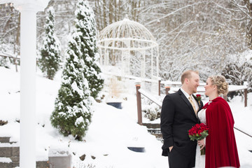 Beautiful bride and groom embracing in snow covered garden wearing red