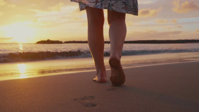 Women's bare feet walk along the sandy beach towards the waves and the beautiful sunset