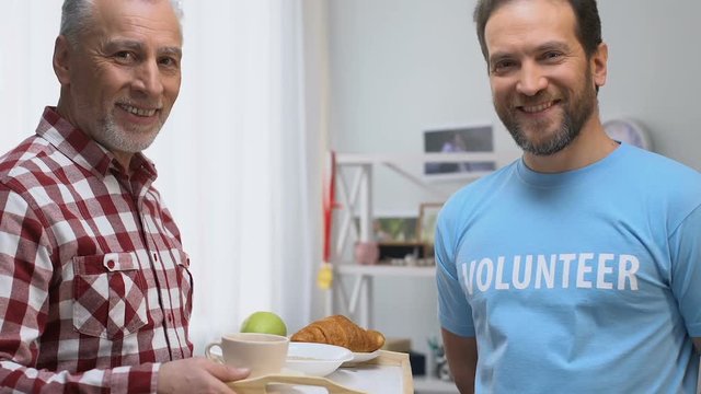 Smiling volunteer bringing breakfast to old man in nursing home care and support