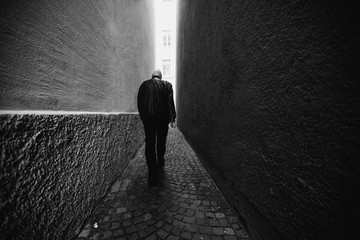 A man in black walking along a narrow alley toward the light. Black and white photography.