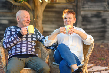 Fototapeta na wymiar Smiling Elderly Man and Woman on Chairs in Outdoor Garden, Vacation. Senior couple relaxing by the park on sunny day.Loving old couple drink coffee.