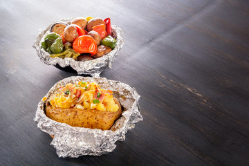 Baked potatoes with bacon, onions and baked vegetables in foil - tomatoes, eggplants, peppers on a gray wooden table.