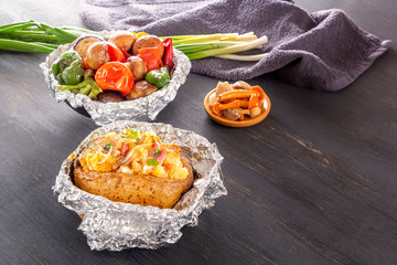 Baked potatoes with bacon, onions and baked vegetables in foil - tomatoes, eggplants, peppers on a gray wooden table.