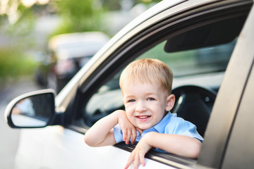 Little blond boy driving a big white car in a blue shirt looks out the window and smiles