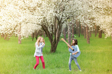 Fototapeta premium kids rock band of two funny little girls with vintage guitar and hairbrush as microphone playing music in spring blossom park outdoor