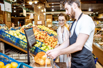 Handsome worker in uniform helping young woman customer to weigh apples on the scales in the supermarket