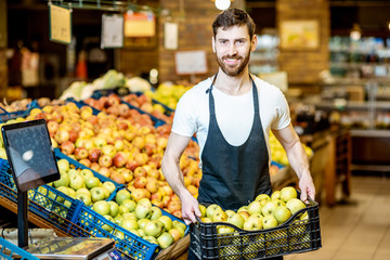 Portrait of a handsome worker or farmer in uniform holding box full of green apples in the shop or supermarket