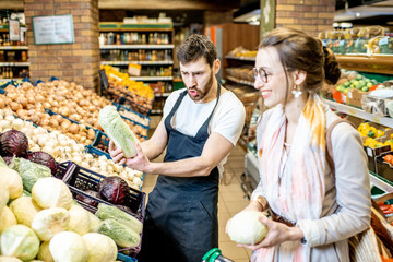 Shop worker helping young woman client to chooose vegetables in the supermarket