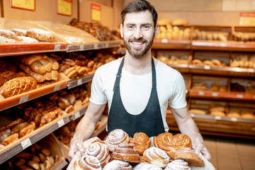 Portrait of a handsome baker in uniform standing with fresh pastries in the bakery deparment of the...