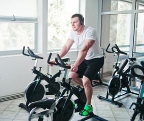Fototapeta na wymiar Healthy lifestyle concept. Young sporty man in white t-shirt and shorts is exercising bike at spinning class . Cardio training