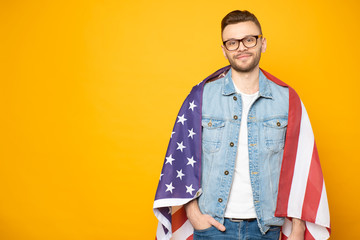 Diligent student. Young handsome guy in denim outfit and matching glasses who studies American English with the three color flag over his shoulders in front of bright yellow background.