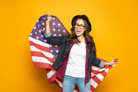 Blowing away. Pleased girl in denim jeans and shirt, white t-shirt and checkered blouse is holding American country symbol and laughing in front of yellow background.