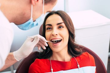 Brilliant smile of a girl in a red sweater sitting in the dentist chair and getting a medical examination of a dentist with a help of dental examination mirror.