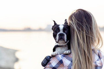 Beautiful girl holding on her hands boston terrier black and white dog on sandy beach near the river