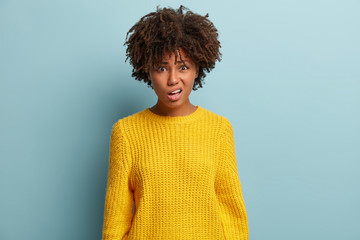 Disappointed angry woman with Afro hairstyle, frowns face, meets rival, displeased to have talk with unpleasant person, wears yellow clothes, isolated over blue wall. People and indignation.