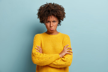Distressed sad dark skinned woman purses lips, looks with jealousy and regret, keeps hands crossed over chest, tells about unfair things, feels offended, wears yellow jumper, isolated on blue wall
