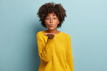 Beautiful woman gives air kiss at camera, has folded lips in mwah, dressed in soft winter wear, isolated over blue background. Love concept. African American girlfriend has lovely expression, gestures