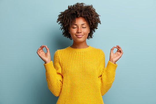 Satisfied dark skinned young woman makes okay gesture with both hands, keeps eyes closed, dressed in yellow clothes, practices yoga after work, isolated over blue studio backround. Body language