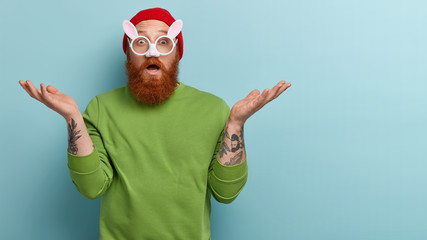Studio shot of astonished puzzled redhead man with thick bristle, wears rabbits glasses with small ears, expresses surprisement, feels uncertain where to celebrate Easter, isolated on blue wall