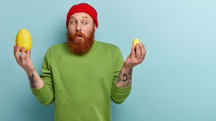 Confused bearded man holds big and little Easter eggs in yellow colour, doesnt know which to choose...