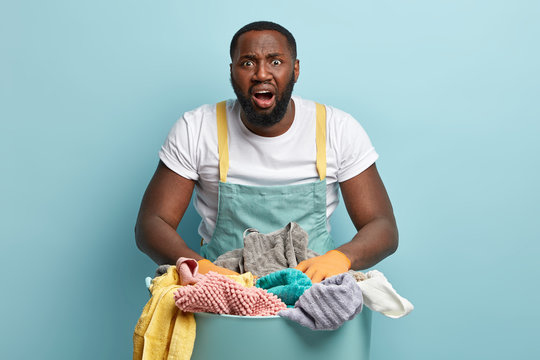 Oh No, Again Washing? Frustrated Black Laundry Hotel Worker Looks At Pile Of Clothes For Ironing, Opens Mouth Widely, Dressed In White T Shirt And Apron, Does Housework And Chores, Stands Indoor