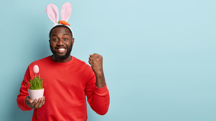 Studio shot of glad Afro American guy holds hand raised in fist, has toothy smile, dressed in red jumper, carries pot with decorated Easter egg, rejoices coming holiday, blank space aside for text