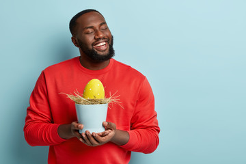 Optimisitic glad Afro American man has dreamy expression, dreams about celebrating Easter day in family circle, holds vase with big yellow egg on hay, shares traditions of religious holiday.