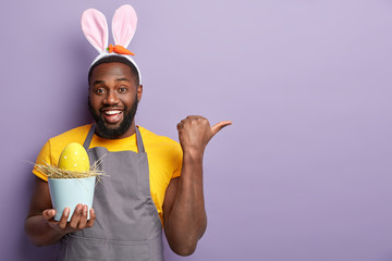 Plump glad Afro American man points away with thumb, glad to advertise something, holds pot containing egg, decorates with colorful paints, isolated over lilac studio wall. Easter traditions concept