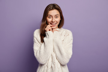 Portrait of attractive lovely woman keeps fore finger on lips, has gentle pleased smile, listens pleasant words of praise, dressed in warm knitted sweater, models over violet wall. Facial expressions