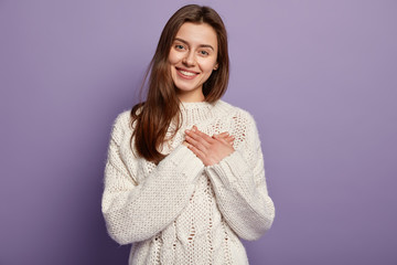 Friendly pleased woman has heart filled with love and gratitude, keeps both hands on chest, has lovely sincere smile, wears oversized jumper, stands against purple background, sympatizes friend