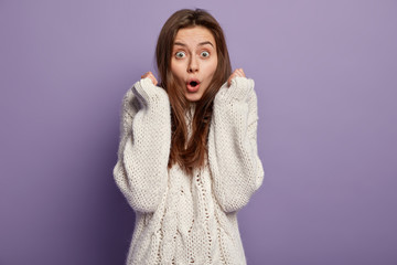 Waist up shot of shocked lovely woman clasps hands, keeps mouth opened, frightened by terrified scene, being speechless, wears long sleeved oversized jumper, stands against purple background