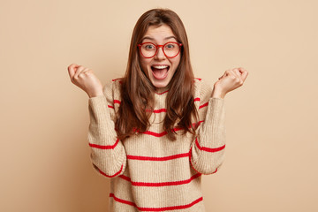 Photo of pleased young woman raises hands, shouts with excitement, wears optical glasses and...
