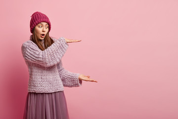 Shocked young European woman makes big gesture, keeps mouth widely opened, wears hat and knitted jumper, shows height of something, isolated over pink background. Thats enough big, I should say