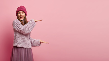 Impressive cheerful woman demonstrates size of present she received, wears headgear, loose sweater and long skirt, gestures over pink background, demonstrates measure sign, big object or thing