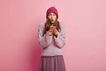 Stupefied lovely woman holds cell phone, receives message with astonishing news, wears headgear, loose sweater and skirt, types feedback, isolated over pink background, surfes social network