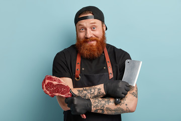 Bearded joyful male chef or cook holds sharp knife for chopping meat, piece of raw fresh pork,...