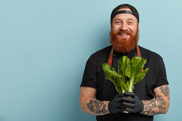 Dieting and healthy eating concept. Cheerful male chef holds green salad, shares recipe of vegetarian dish, supports healthy ration, says eat vegetables which contain vitamins, likes cooking process - obrazy, fototapety, plakaty