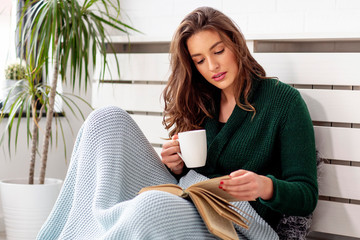 Girl with coffee mug in hand reading book wrapped in blanket