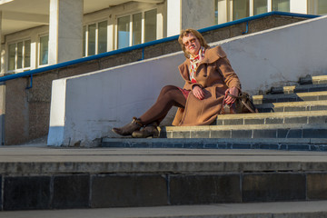 Attractive middle-aged woman wearing stylish coat and shoes sitting with bag on stairs step of office building in early spring at sunset. Street city portrait, fashion urban outfit
