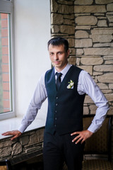 The groom stands near a large window and waits for his bride