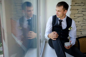 A handsome man sits on the windowsill of a large window and drinks his morning coffee