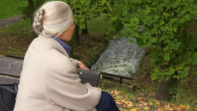 An elderly woman sits on a bench in a park and looks at a creek below