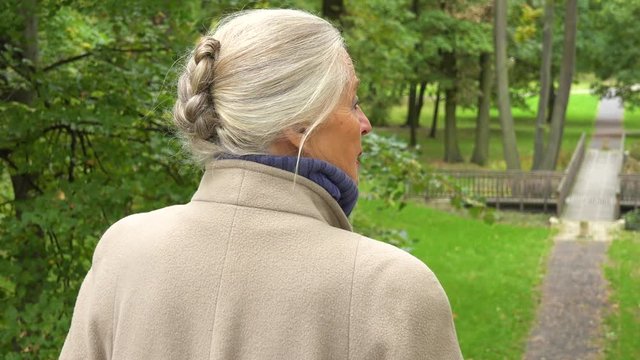 An elderly woman stands in a park and looks around - a footbridge in the blurry background