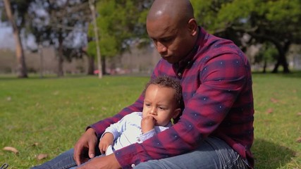 Afro-american young bald father in striped shirt and jeans sitting on grass in park, holding his little mixed-race son on knees, hugging. Family, holiday concept