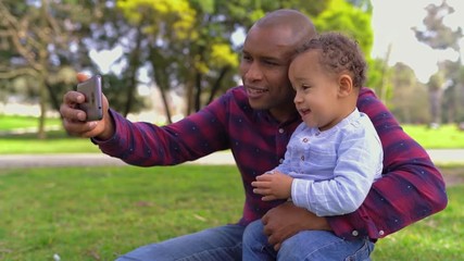 Afro-american young bald father in striped shirt and jeans sitting on haunches in park, holding his little mixed-race son on knees, making selfie. Side view. Family, holiday concept