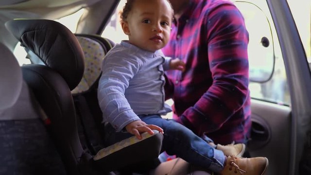 Afro-american young bald father in striped shirt and jeans putting his little mixed-race boy into baby seat, fastening seat belts. Side view. Family, safety concept 