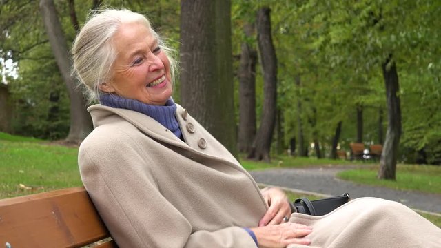 An elderly woman sits on a bench in a park and laughs