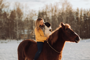 Young happy cute smiling woman with her dog border collie sit on horse in snow field on sunset. yrllow dress